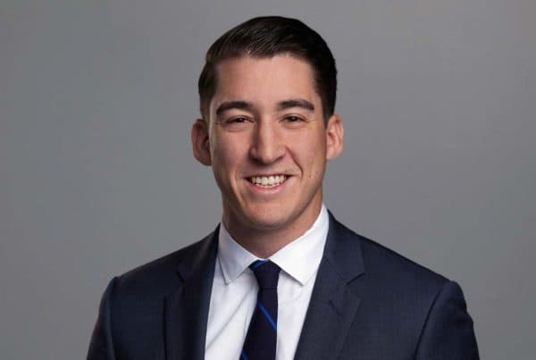Baker J Professional Business Portrait Professional headshot of Baker J smiling in a navy suit with white shirt and blue striped tie against gray background