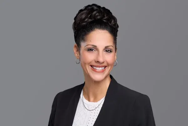 Allison Floyd Professional Business Portrait Professional headshot of Allison Floyd smiling, wearing a black blazer and white top against a gray background.