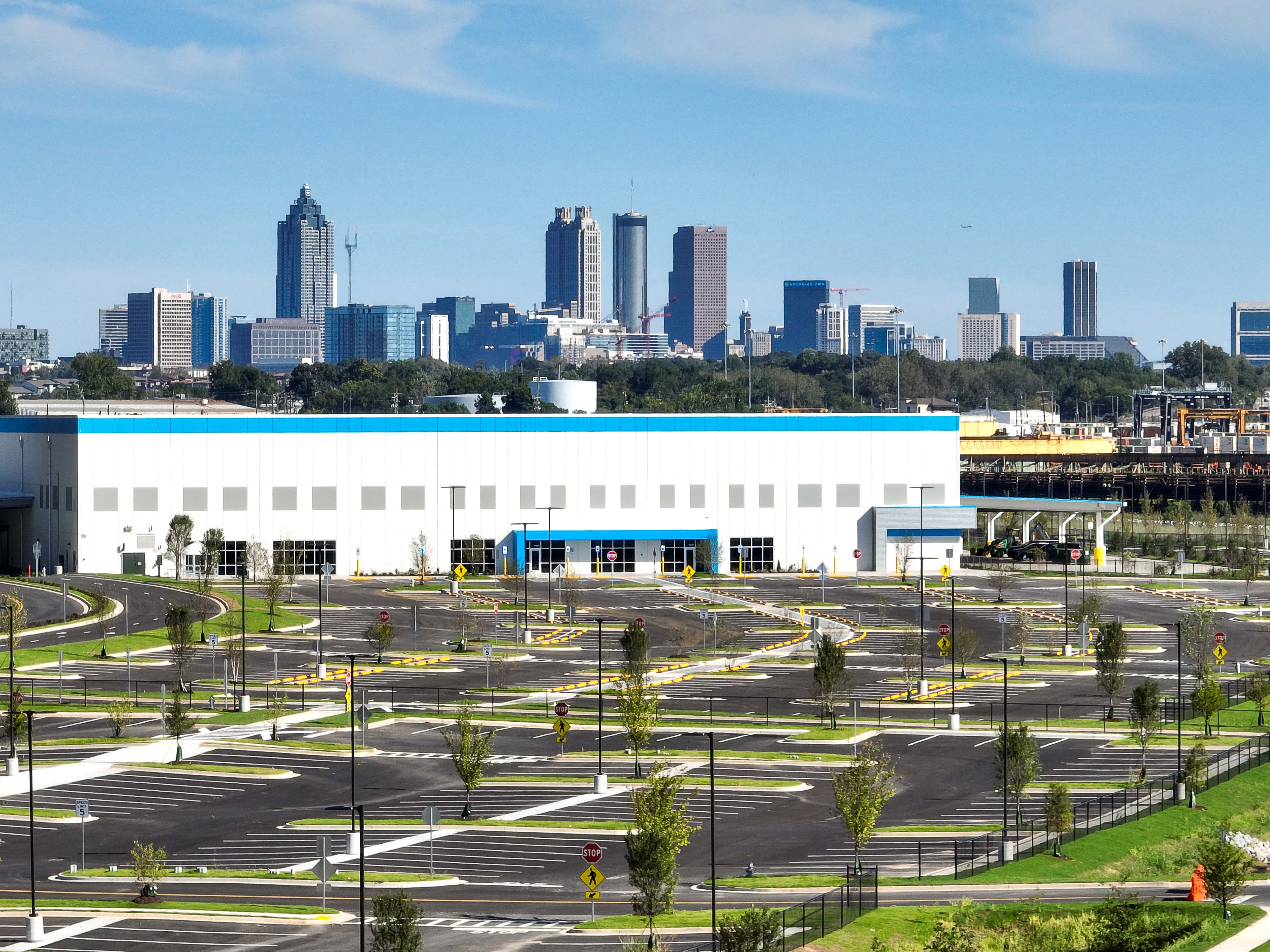 Modern Commercial Warehouse Building with City Skyline View Large white industrial warehouse with blue accents in front of urban skyline, surrounded by new parking lot and landscaping.