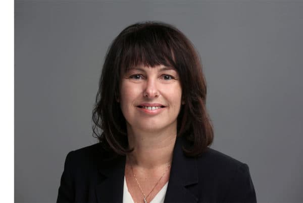 Alexandra Logan Professional Portrait Photograph Professional headshot of Alexandra Logan, a woman with shoulder-length dark hair wearing a black blazer, smiling at camera against gray background.