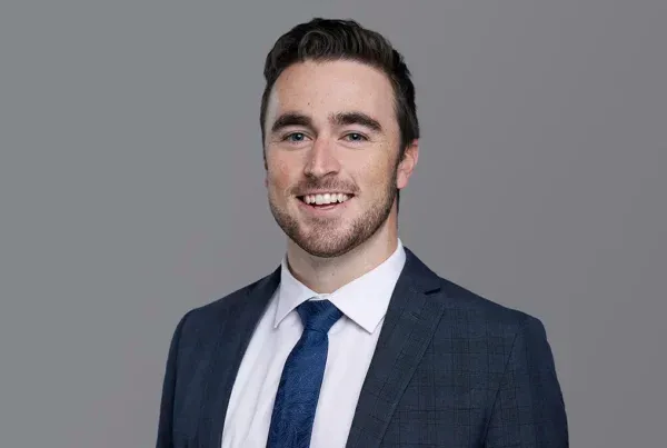 Professional Headshot of Nick in Business Attire Professional headshot of a smiling man named Nick wearing a navy suit, white shirt, and blue tie against a gray background.